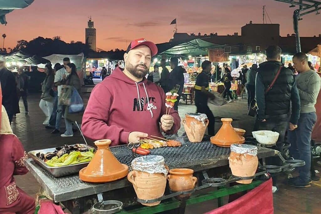 Street-food vendor grilling brochettes in Jemaa el-Fnaa with Koutoubia Mosque at sunset