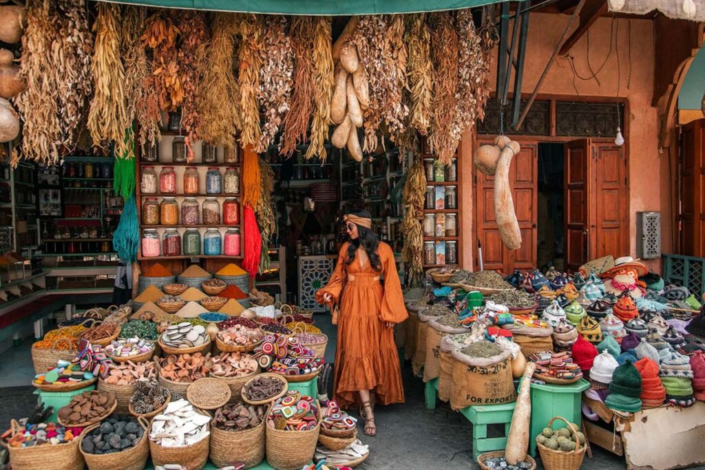 Woman browsing colourful spice stall in Marrakech souk packed with jars, herbs & baskets