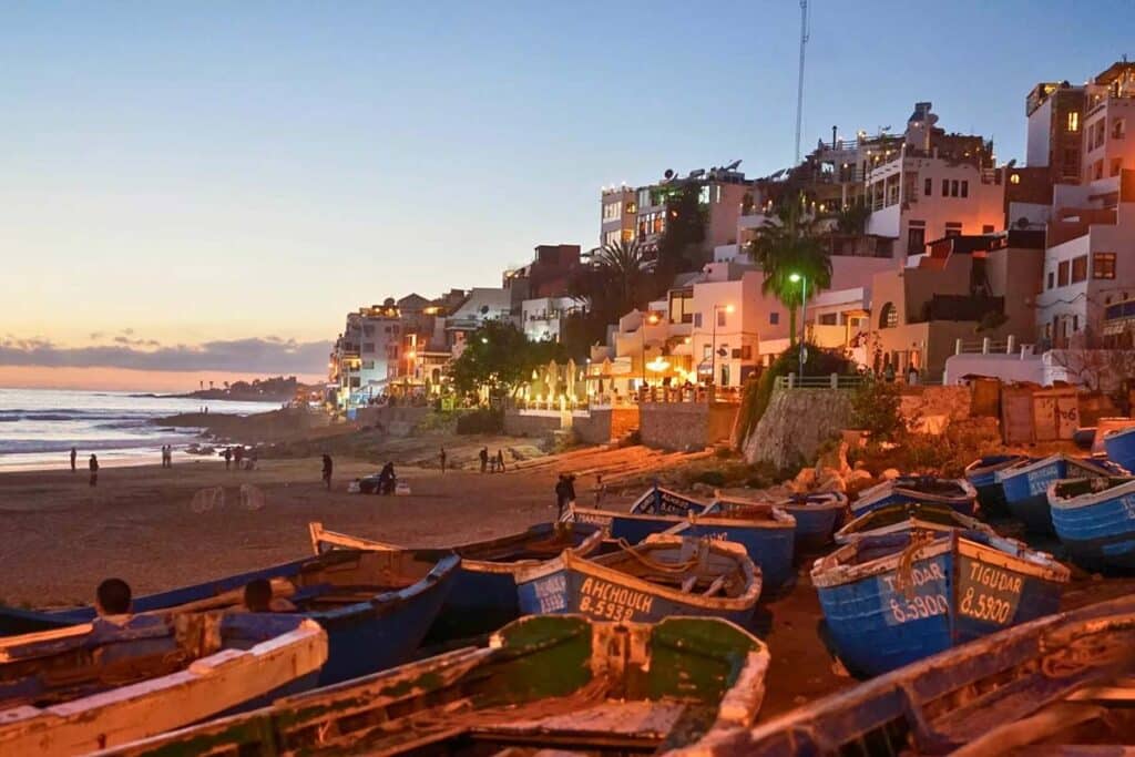 Colourful fishing boats on Taghazout beach at dusk with village lights along Morocco’s Atlantic Coast